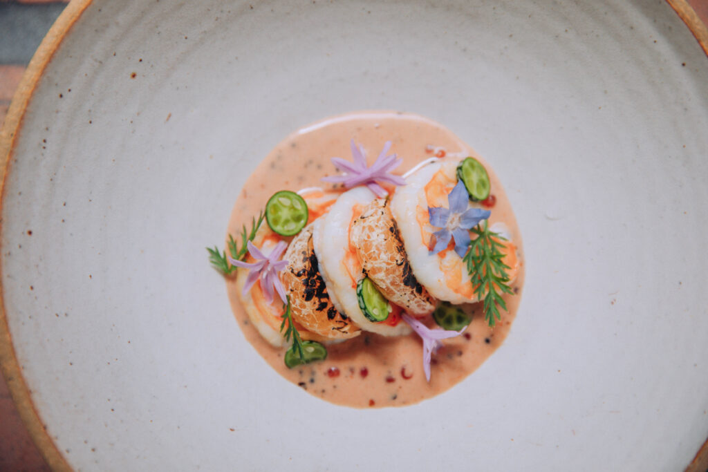 A bowl with three cooked prawns sitting in sauce and garnished with flowers and small vegetables.